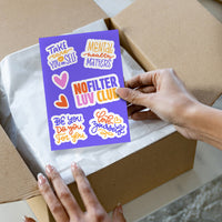 A hand holds a purple sticker sheet with colorful motivational quotes and hearts over an open cardboard box with white tissue paper.