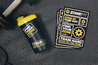 A black shaker bottle with a yellow lid and matching black-and-yellow ZoneFit stickers are arranged on a dark textured surface next to a dumbbell. The stickers read 'ZONEFIT', 'FIND YOUR ZONE', 'TRAIN HARD', and 'STAY IN THE ZONE'.