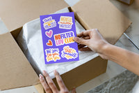 A hand holds a purple sticker sheet with colorful retro-style text stickers over an open cardboard box with white tissue paper.

