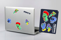 A silver laptop and a black sticker sheet featuring colorful cartoon stickers, including a green zombie-like sock, blue skulls, a slice of pizza, and mushrooms. The laptop has several stickers applied, and the sheet displays the full set of designs.
